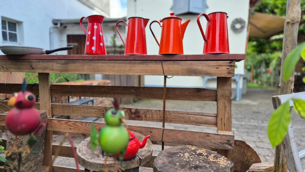 4 rote Emaille Kaffeekannen auf einer senkrechten zweckenfremdeterer Pallette im vordergund ein Blechvogel.