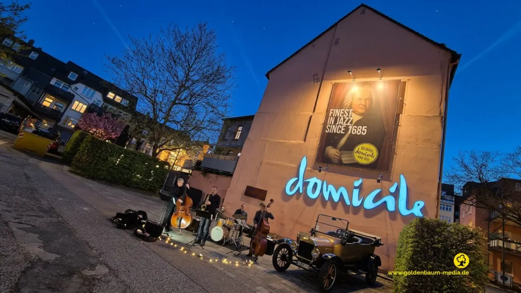 Fassade vom Jazzclub Domicile in Pforzheim Brötzingen. Auf dem Parkplatz steht ein Oldtimer, daneben spielt eine Jazzband