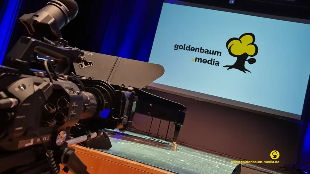 Profesionelle Filmkamera seitlich angeschnitten zeigt eine leinwand in einem Sall, auf der das goldenbaum-Media Logo eingeblendet ost