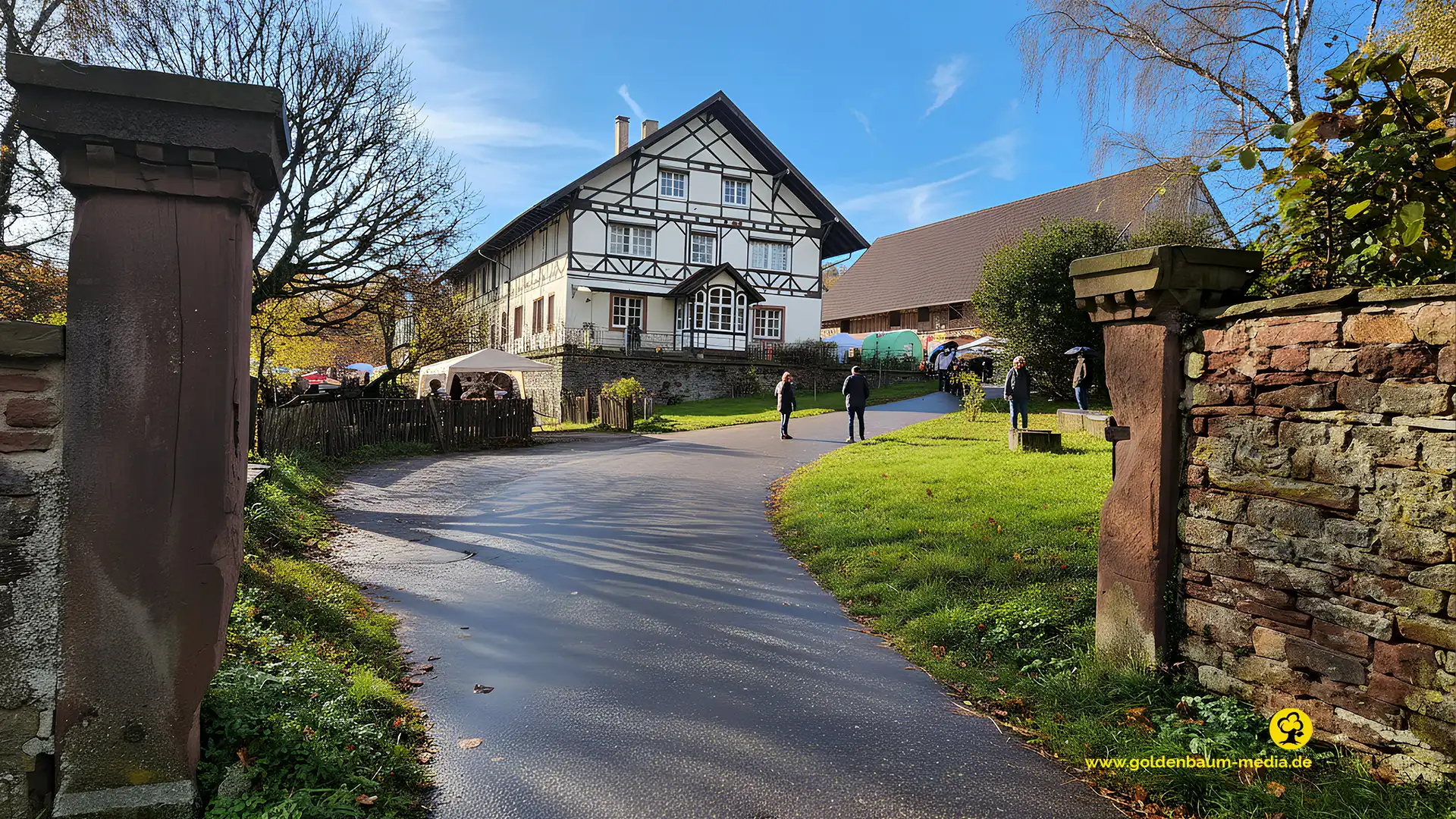 Ein Weg für durch ein Eingangsprotal mit 2 seitlichen Steinpfosten aus Buntsandstein. Im Hintergrund eröffnet sich ein hitorisches Hofgut mit Fachwerkhaus.
